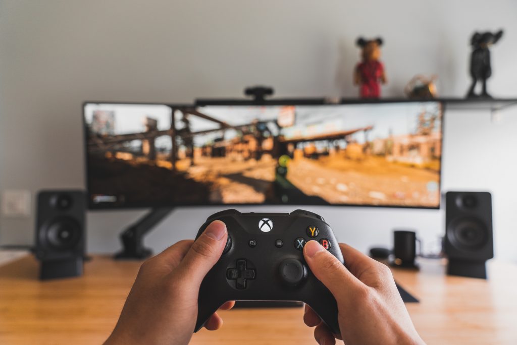 Mãos segurando controle de vídeo game e a melhor caixa de som para PC ao fundo